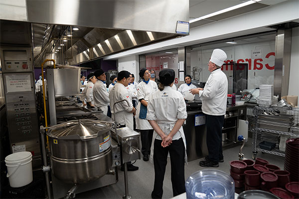 large group of students gathered around a kitchen listening to a teacher