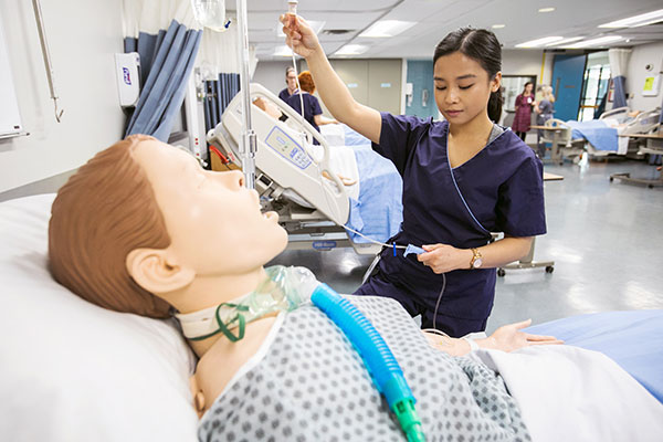 A nurse in navy scrubs adjusts an IV drip on a medical mannequin in a simulation lab
