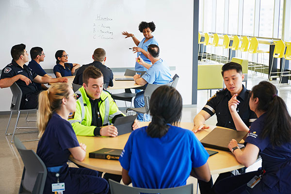A diverse group of medical professionals in uniforms sit around tables, attentively listening to a presenter